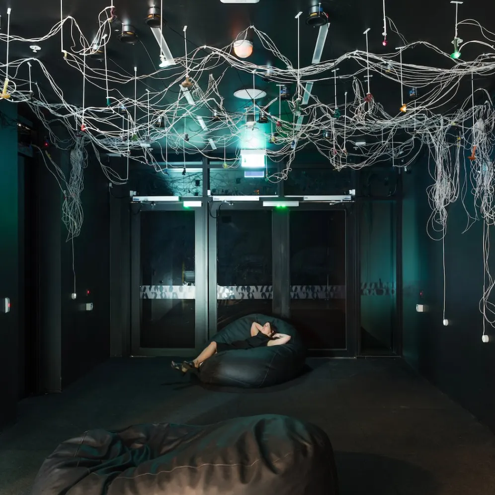 A person lies on a black bean bag in a dark room with hanging wires and light bulbs on the ceiling. The space appears modern and minimalistic, with large glass doors at the back and another large bean bag in the foreground.