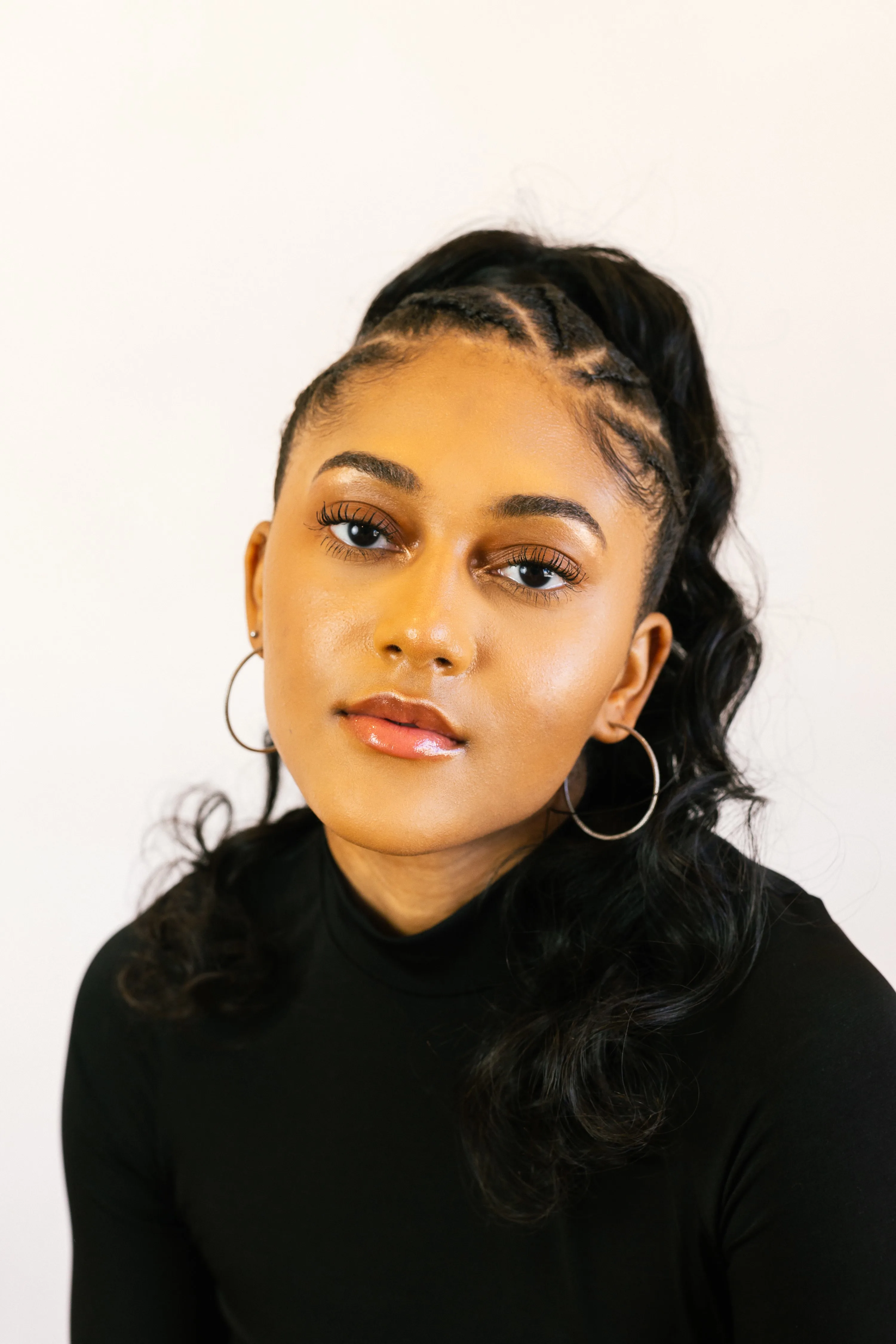 A young woman with long, wavy black hair styled in a high ponytail, wearing hoop earrings and a black turtleneck, looks calmly at the camera against a plain white background.