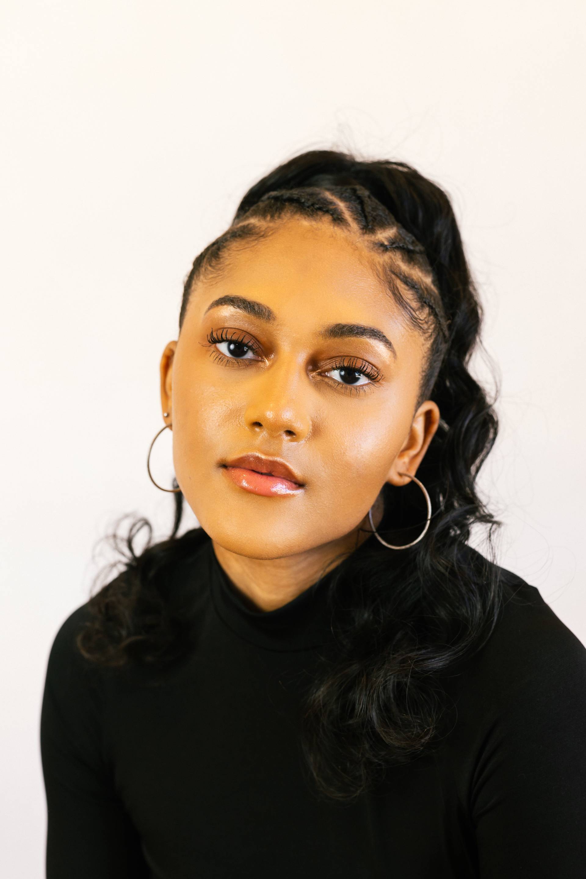 A young woman with long, wavy black hair styled in a high ponytail, wearing hoop earrings and a black turtleneck, looks calmly at the camera against a plain white background.