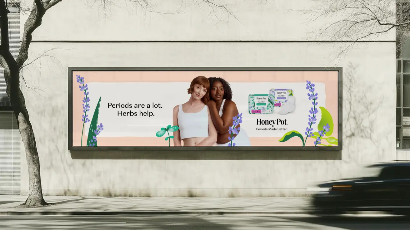 A billboard ad shows two women embracing, one with fair skin and red hair, the other with dark skin and curly hair. Text reads: Periods are a lot. Herbs help. and features Honey Pot period products with floral illustrations.