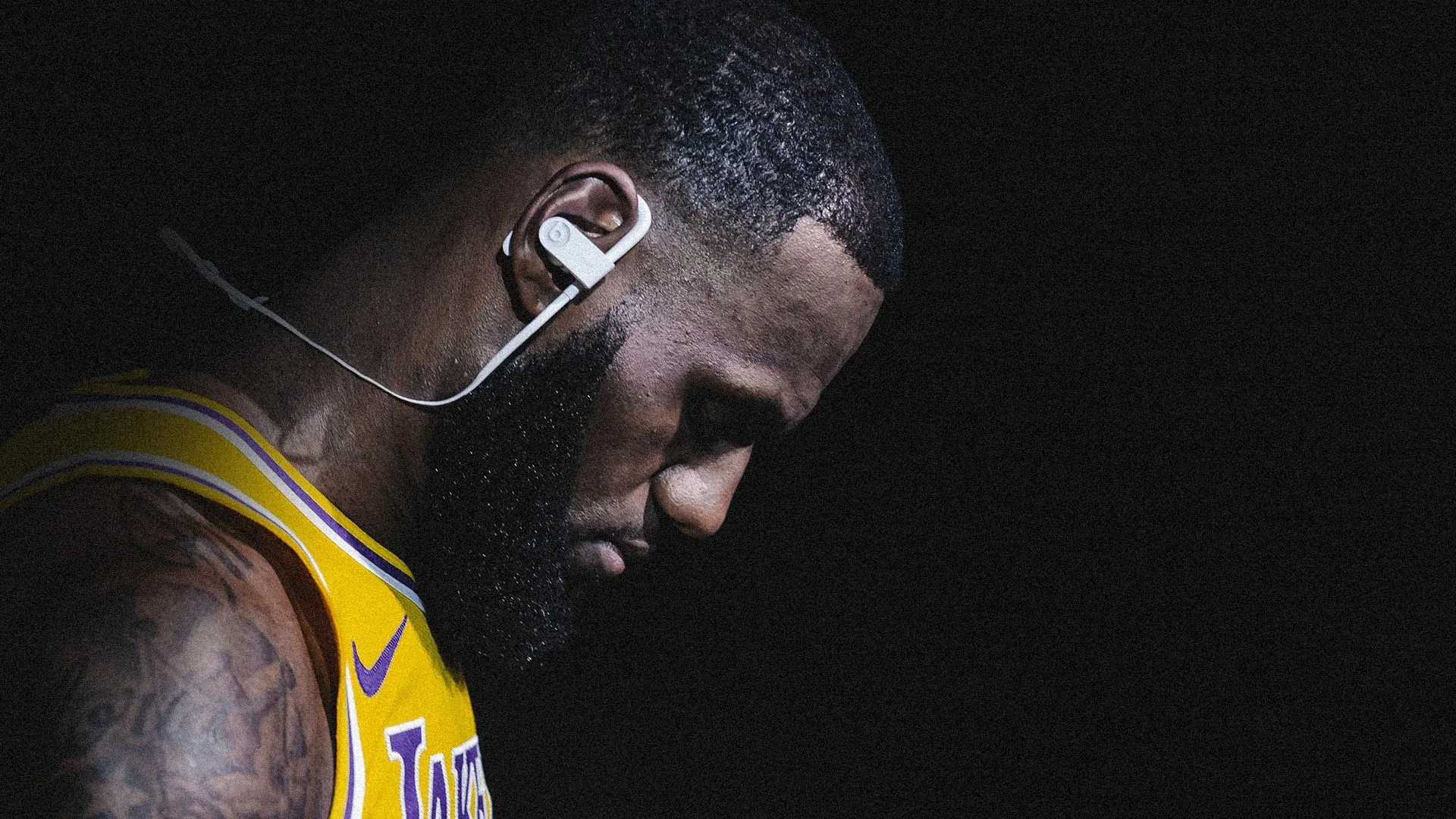 A basketball player wearing a yellow jersey and wireless earphones looks down in a dark, dramatic setting, with light highlighting his face and upper body.
