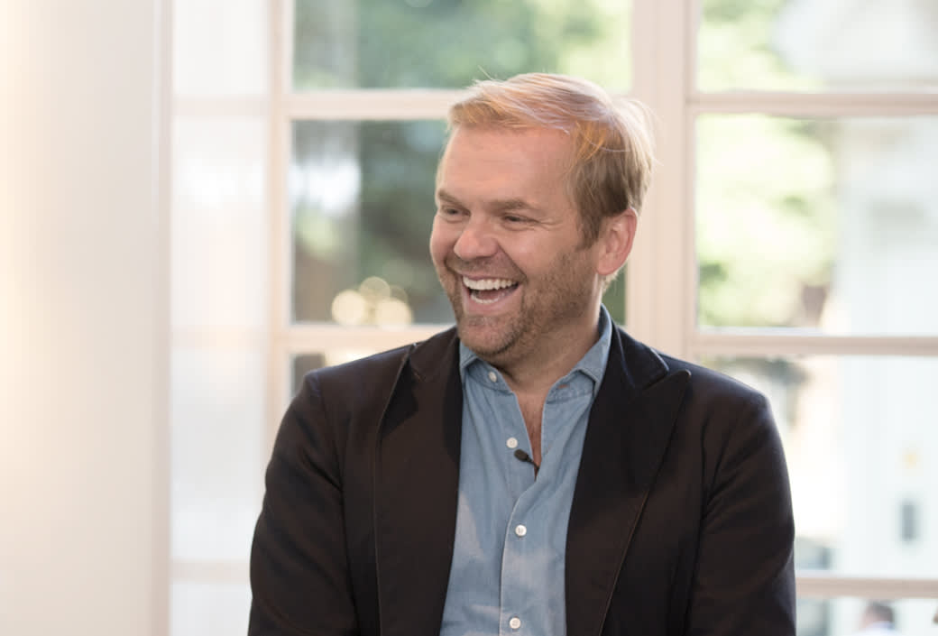 A man with light hair and a beard, wearing a blue shirt and dark blazer, sits smiling and laughing in front of large windows with greenery visible outside.
