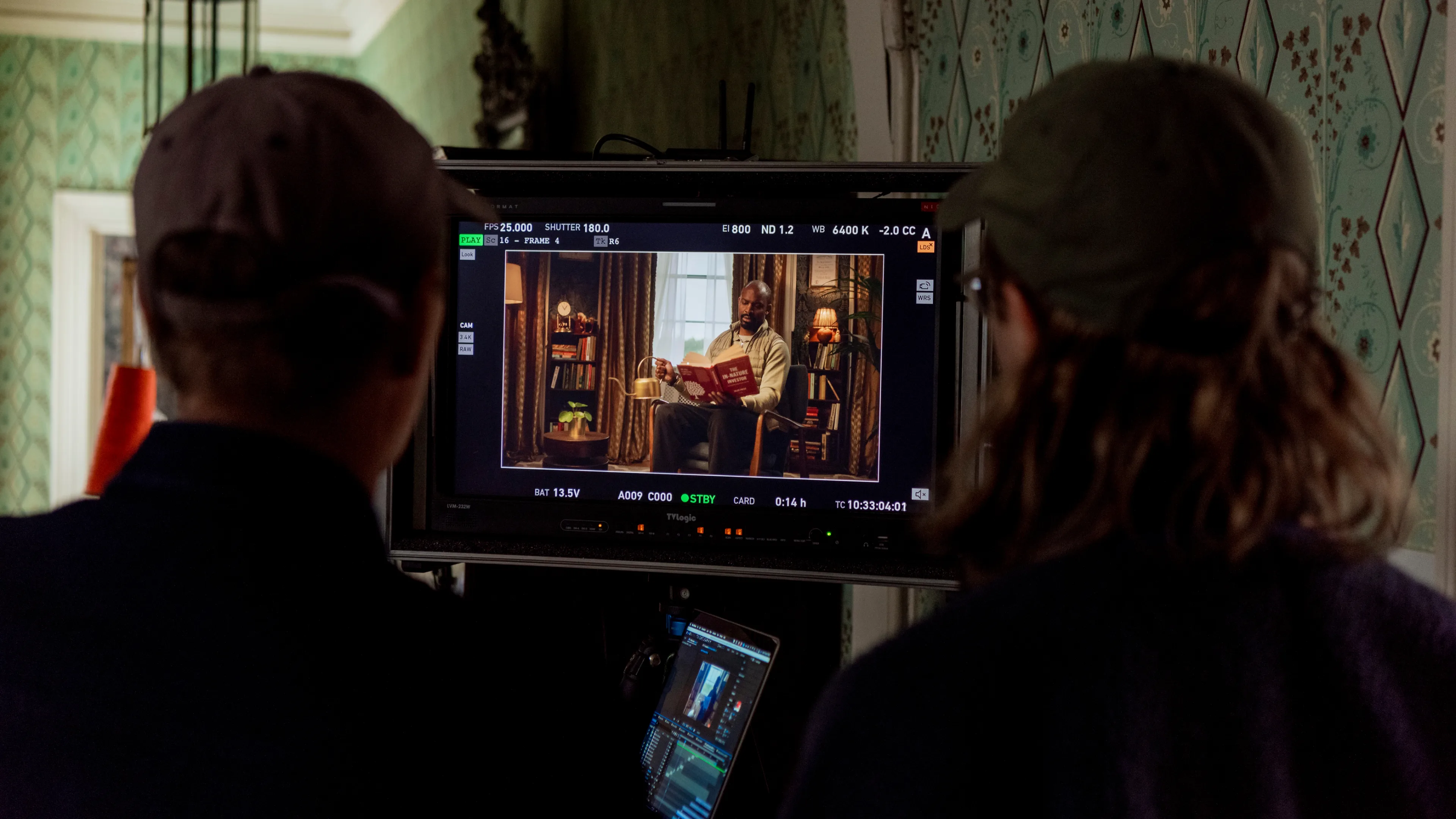 Two people watch a monitor displaying a scene being filmed, showing a person reading a book seated in a warmly lit, cozy room with shelves and decor in the background.