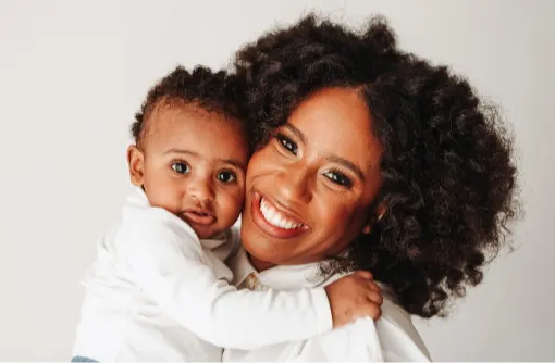 A woman with curly hair smiles brightly while holding a young child, who has short curly hair and looks at the camera. Both are wearing white tops and are set against a plain light background.
