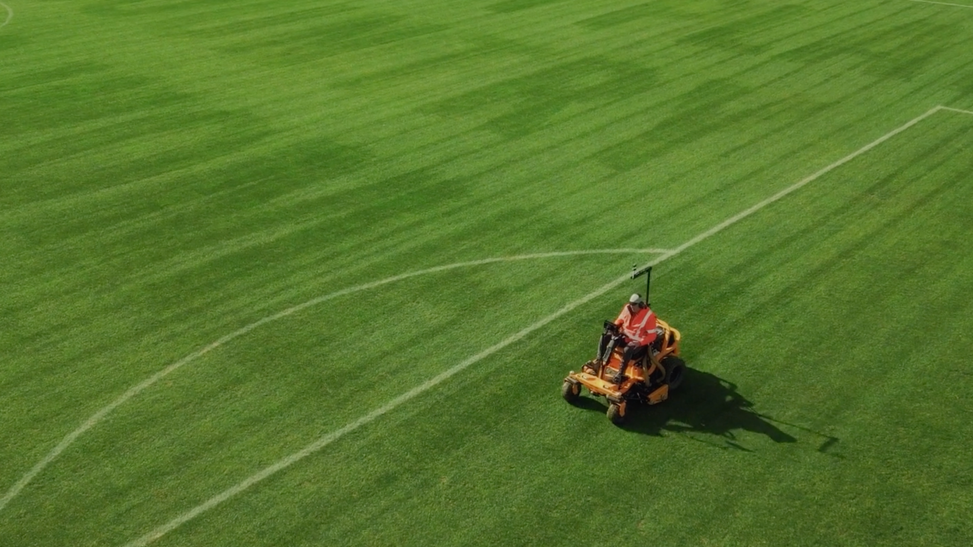 A ride-on lawnmower rides across a football pitch with a driver seated, faint lettering is seen in the grass.