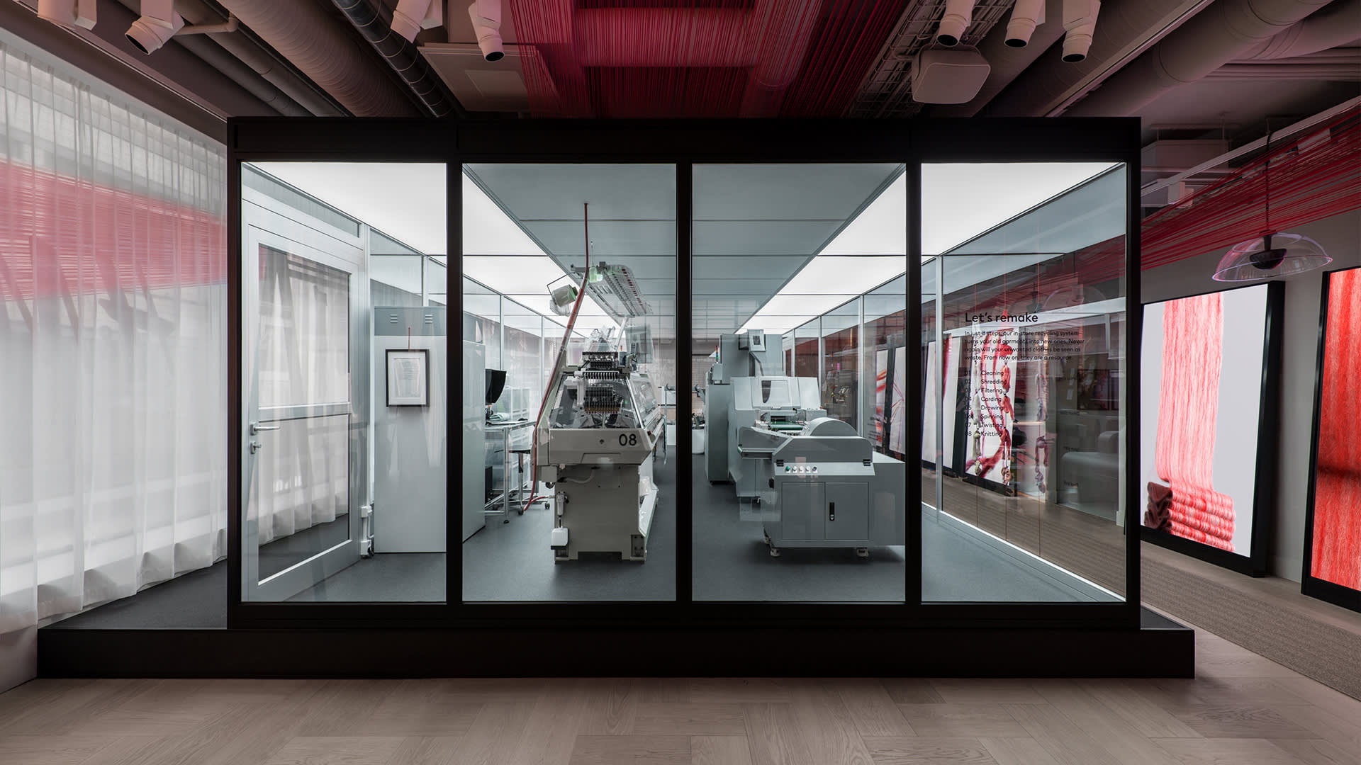 A large glass display case contains modern industrial textile machines in a well-lit showroom, surrounded by red and white fabric panels and ceiling pipes.