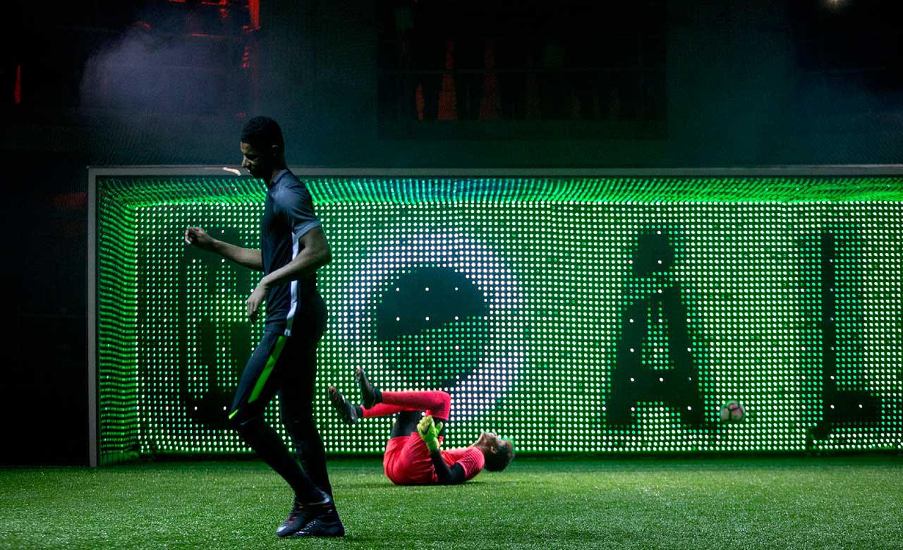 A soccer player in black celebrates in front of a lit-up GOAL sign, while a goalkeeper in red lies on the ground inside the net.
