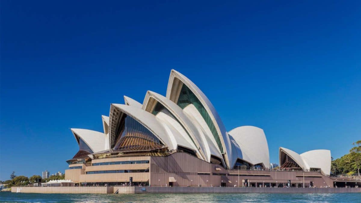The Sydney Opera House with its iconic white sail-like roof structures against a clear blue sky, viewed from across the water during daylight.