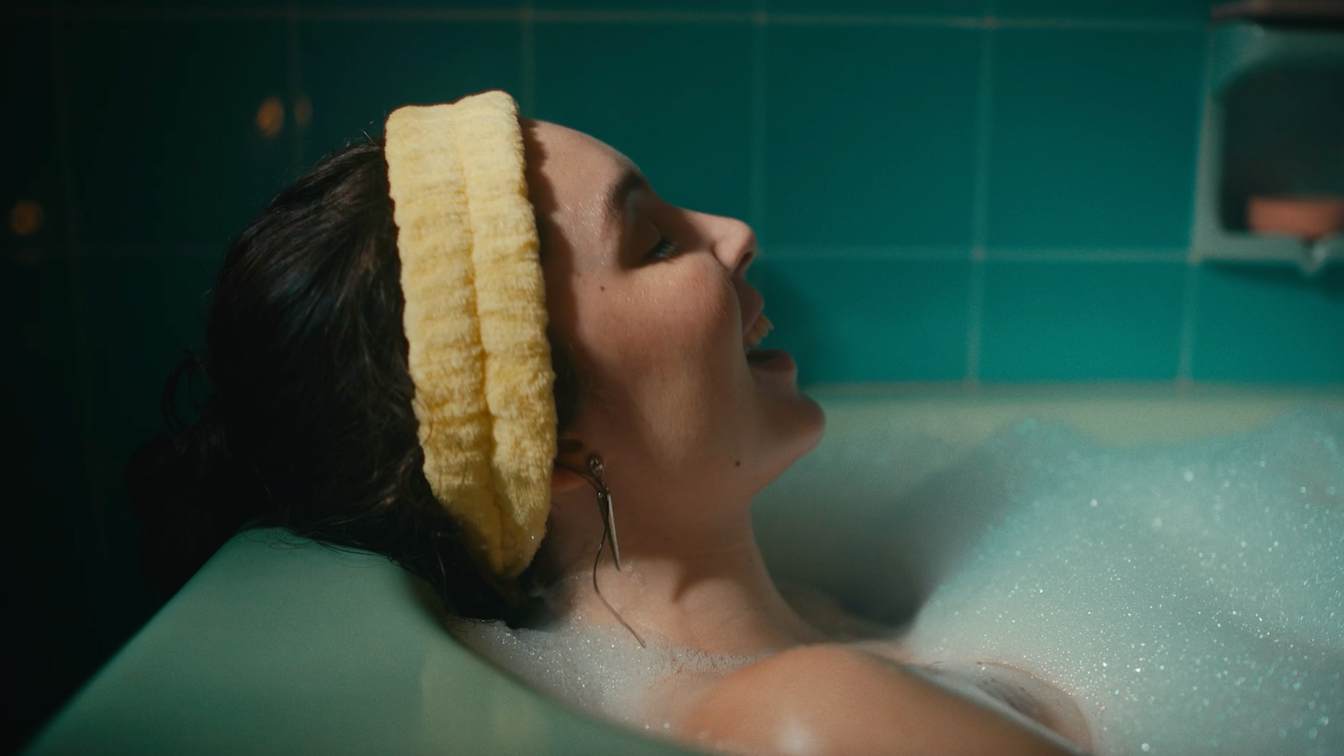 A woman with a yellow headband relaxes in a bubble-filled bathtub, eyes closed and smiling, against a backdrop of teal bathroom tiles.