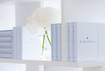A white shelf displays several copies of a book titled Limitless with light blue covers. In the background, there is a glass vase holding white flowers. The scene has a clean, minimalist aesthetic.