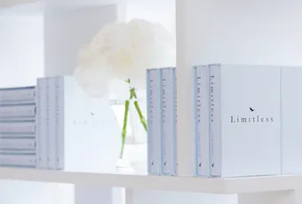 A white shelf displays several copies of a book titled Limitless with light blue covers. In the background, there is a glass vase holding white flowers. The scene has a clean, minimalist aesthetic.