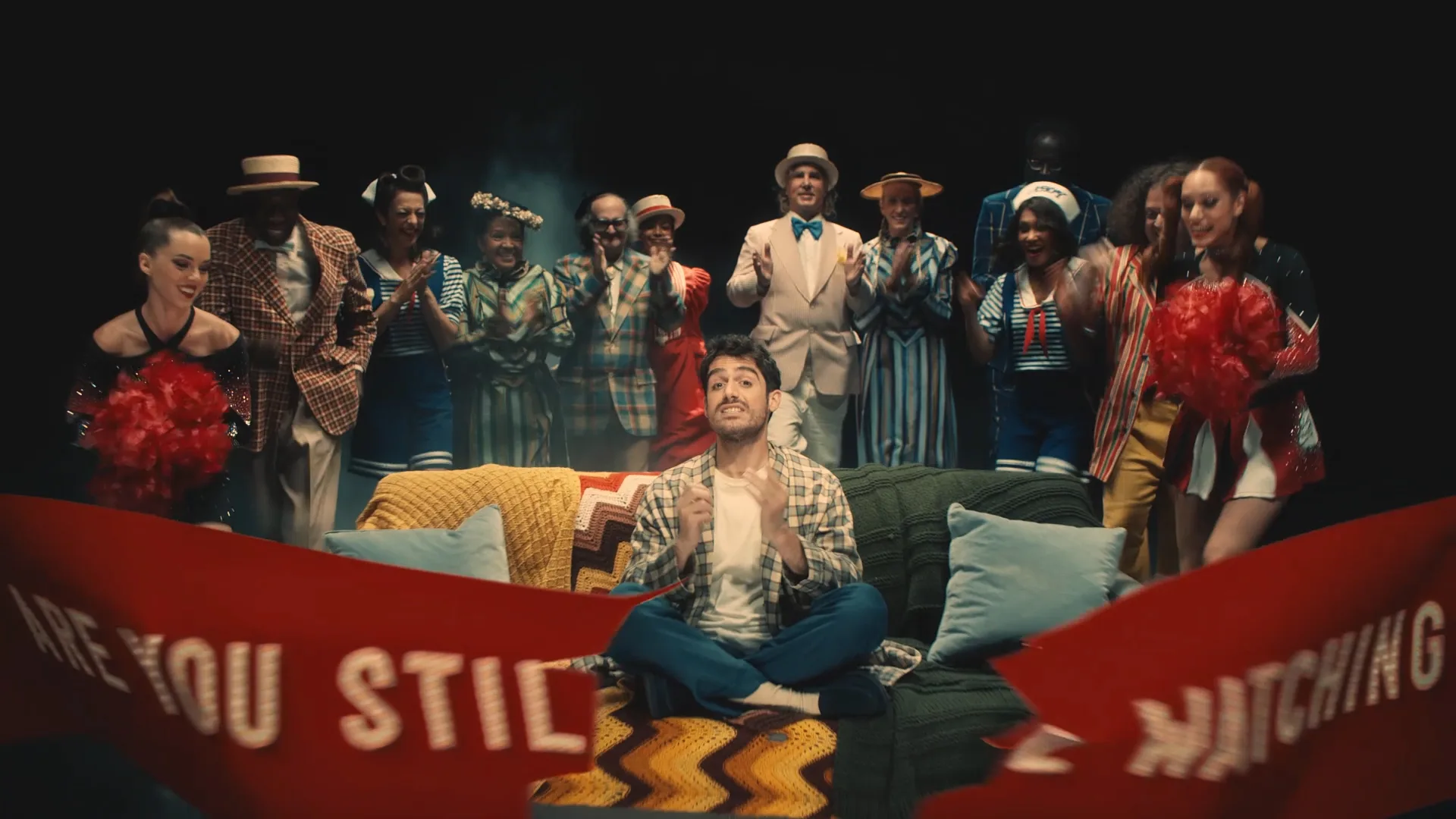 A man sits on a couch looking startled as a group of lively people in colorful costumes and retro outfits cheer and clap behind him. A red banner in front reads, ARE YOU STILL WATCHING?.