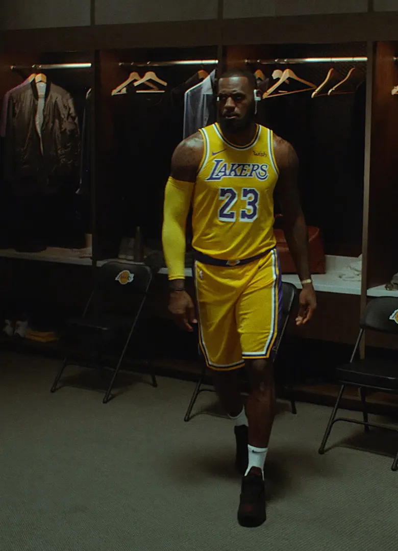 A basketball player in a yellow Los Angeles Lakers uniform with the number 23 walks in a locker room. Jackets and empty hangers are visible in the lockers behind him.