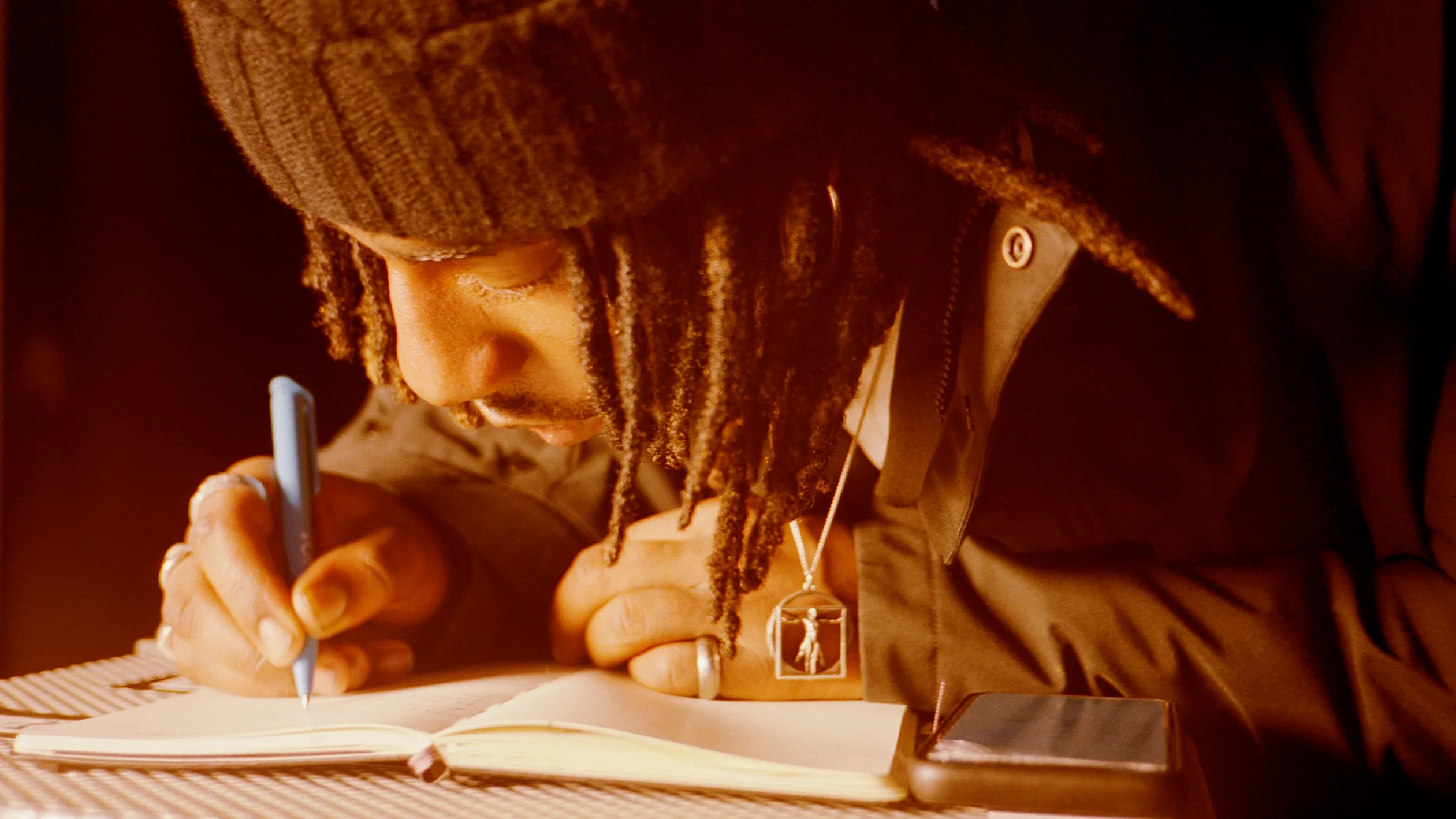 A person with long braided hair, wearing a knit hat and a pendant necklace, writes in a notebook with a blue pen. A smartphone rests on the table beside them. Warm lighting highlights their focus.