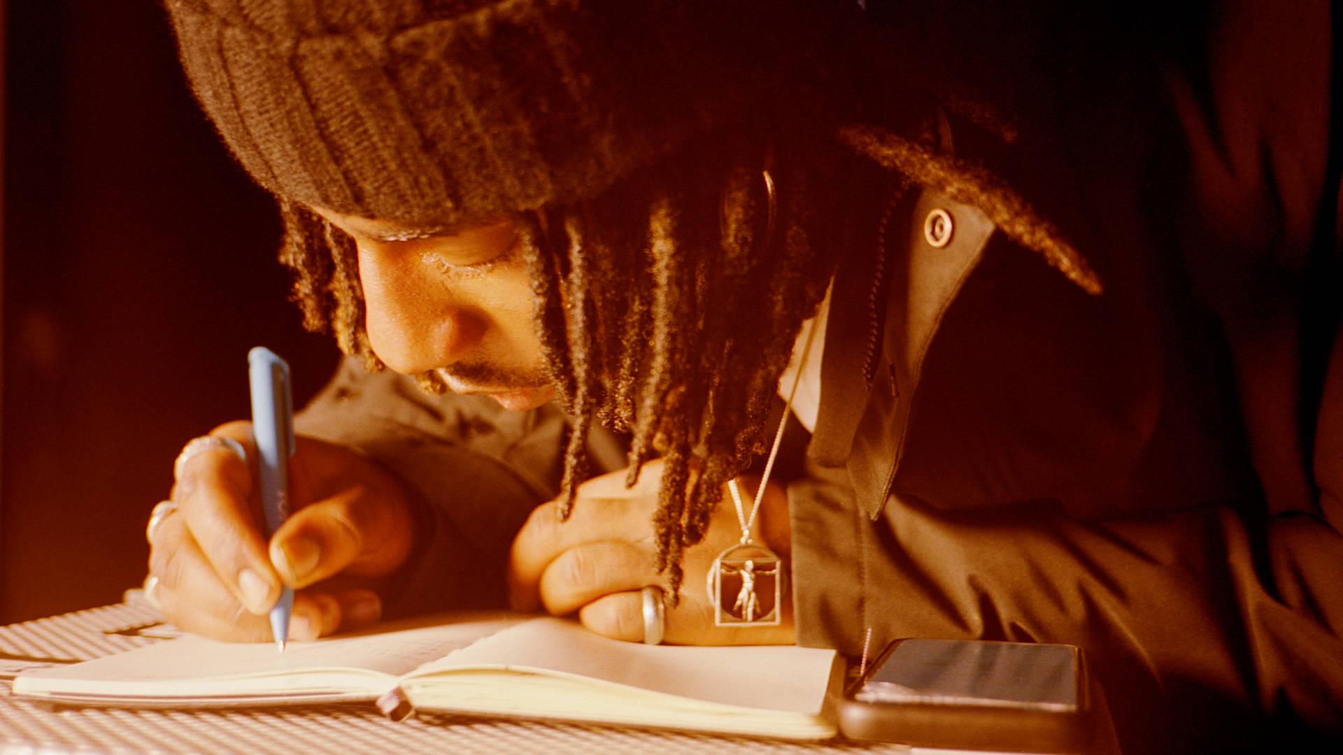 A person with long braided hair, wearing a knit hat and a pendant necklace, writes in a notebook with a blue pen. A smartphone rests on the table beside them. Warm lighting highlights their focus.