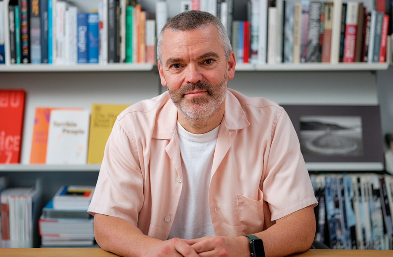 A man with short gray hair and a beard sits at a table, wearing a light pink shirt over a white t-shirt. Bookshelves filled with books are visible in the background.