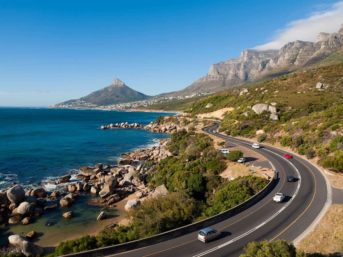 A scenic coastal road curves along the ocean, bordered by rocky shores and lush green hills, with cars driving and mountains rising in the background under a clear blue sky.