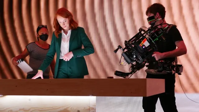 A woman in a green suit stands at a desk under studio lights, while a camera operator films her and another crew member stands nearby, both wearing face masks. The background features a curved, textured wall.