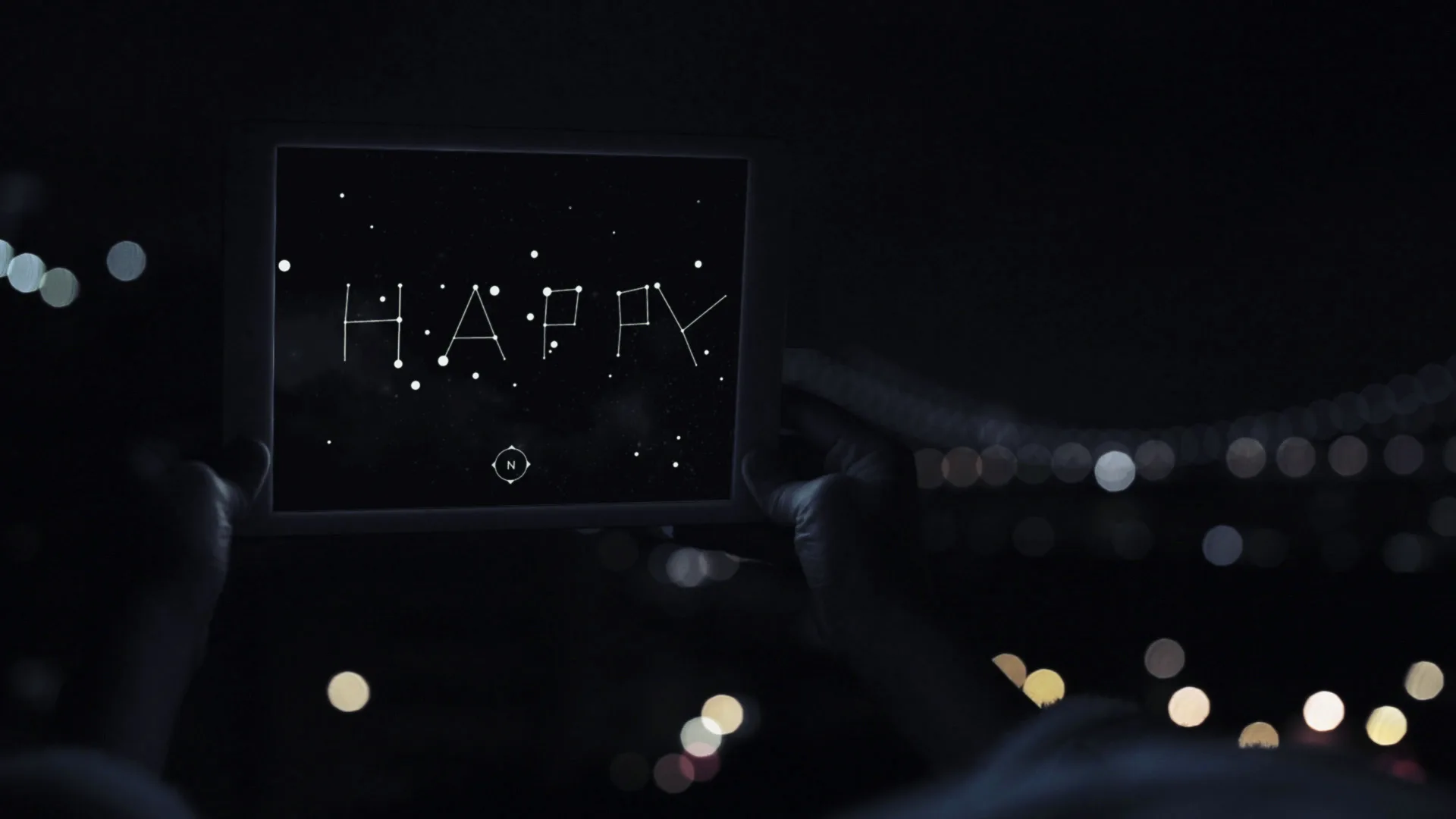 A person holds up a tablet at night, displaying the word HAPPY written in star-shaped dots, resembling constellations, against a dark, starry background. City lights are blurred in the distance.