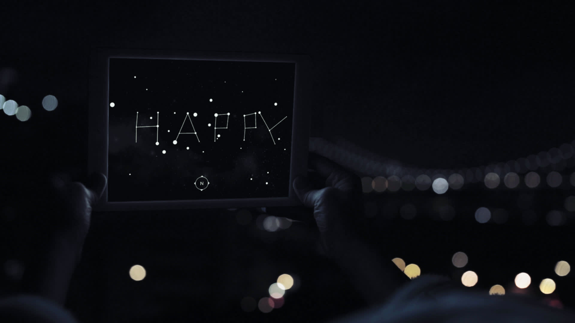 A person holds up a tablet at night, displaying the word HAPPY written in star-shaped dots, resembling constellations, against a dark, starry background. City lights are blurred in the distance.