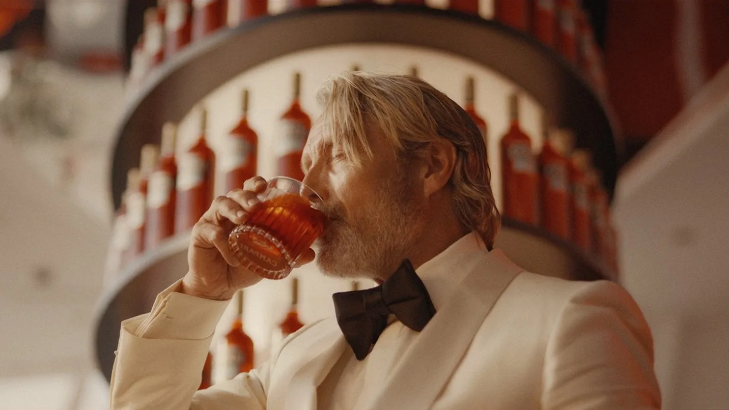 A man in a white tuxedo with a black bow tie drinks from a glass, standing in front of shelves lined with red bottles.