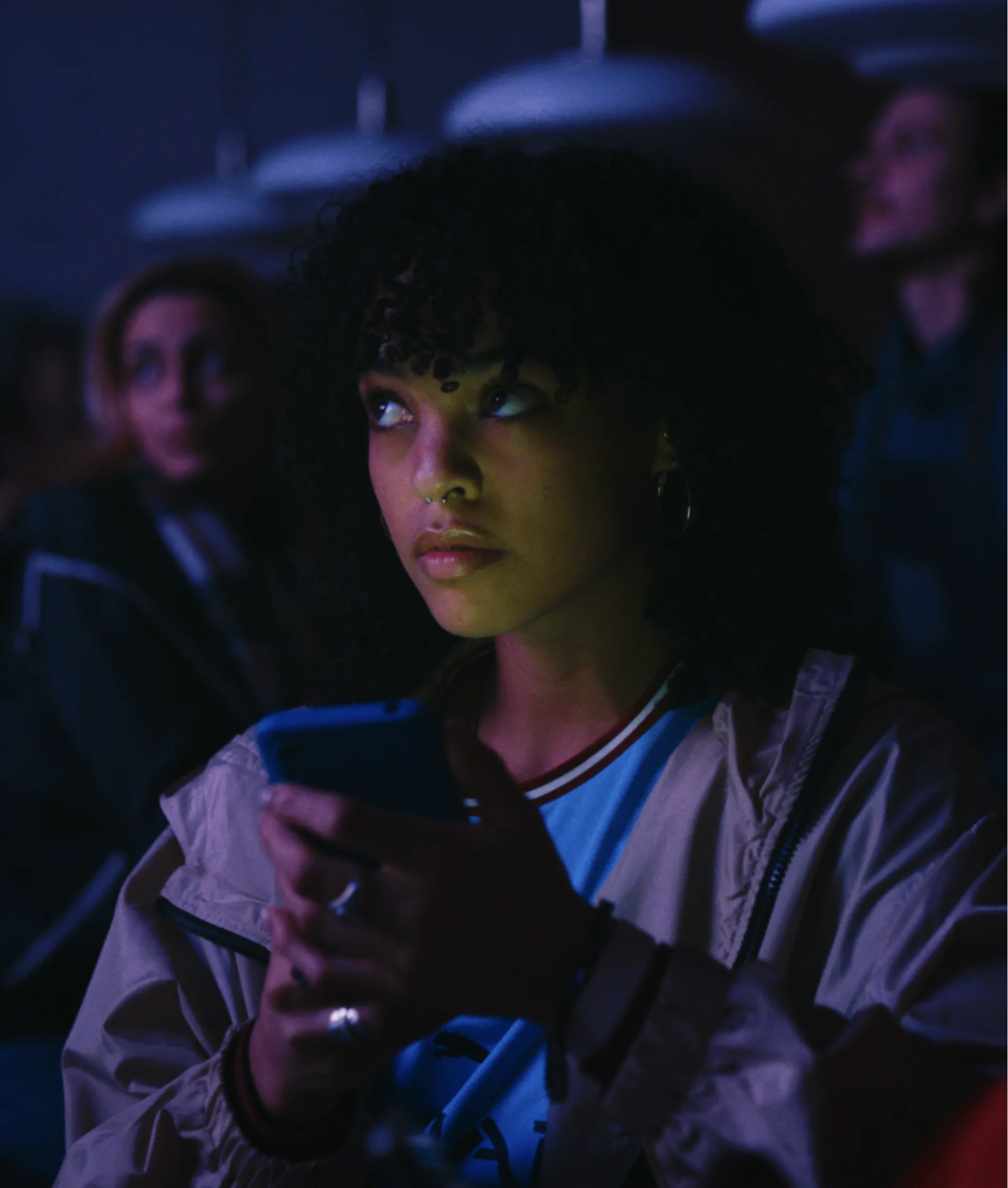 A young woman with curly hair holds a phone and looks up thoughtfully in a dimly lit room, with other people blurred in the background.