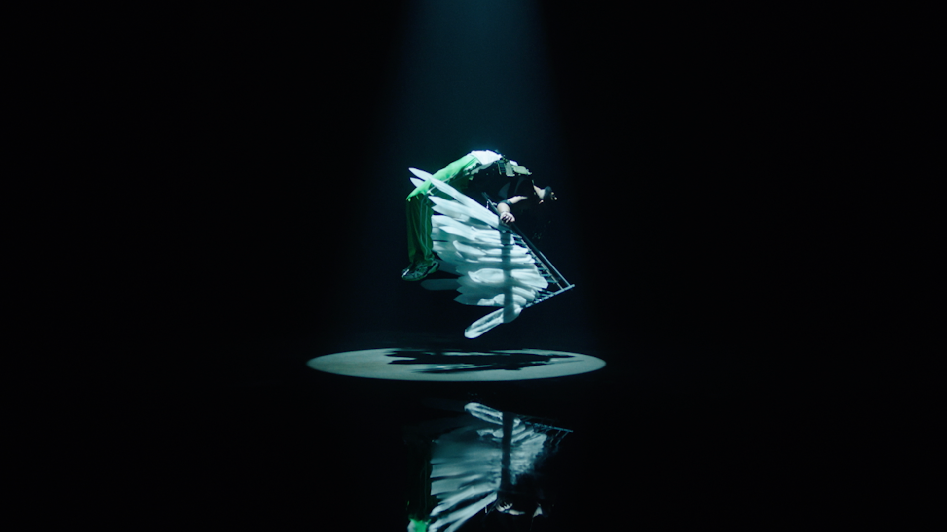 A person dressed in green and black with large white feathers is suspended midair under a single spotlight in a dark room, casting a dramatic shadow on the floor below.