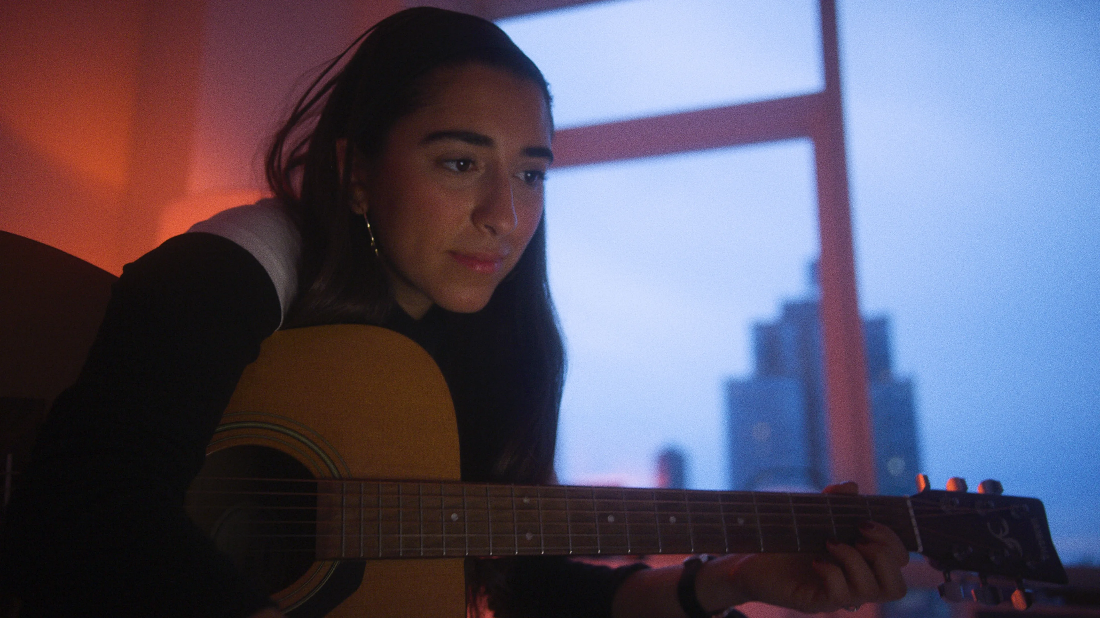A woman with long dark hair plays an acoustic guitar indoors by a window at dusk, with city buildings visible outside and warm ambient lighting inside.