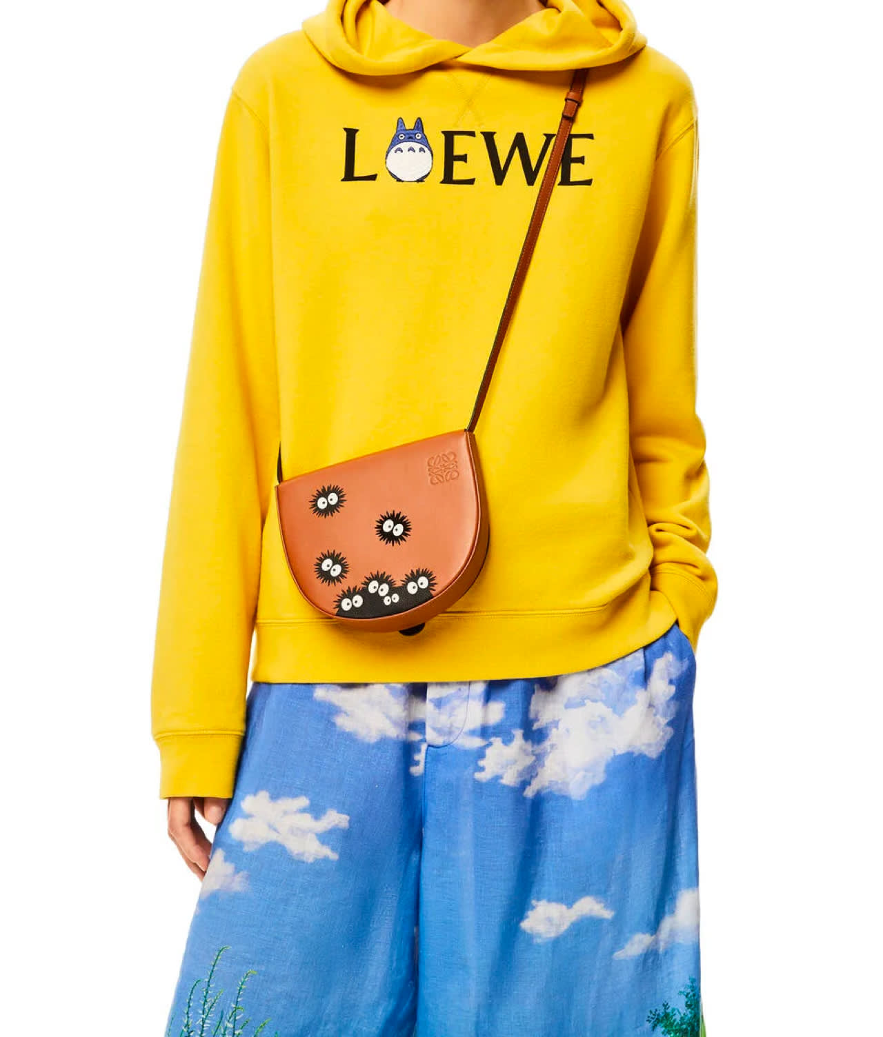 A person wearing a bright yellow Loewe hoodie with a cartoon character, blue pants with a cloud pattern, and carrying a tan crossbody bag decorated with small black and white cartoon soot sprites.