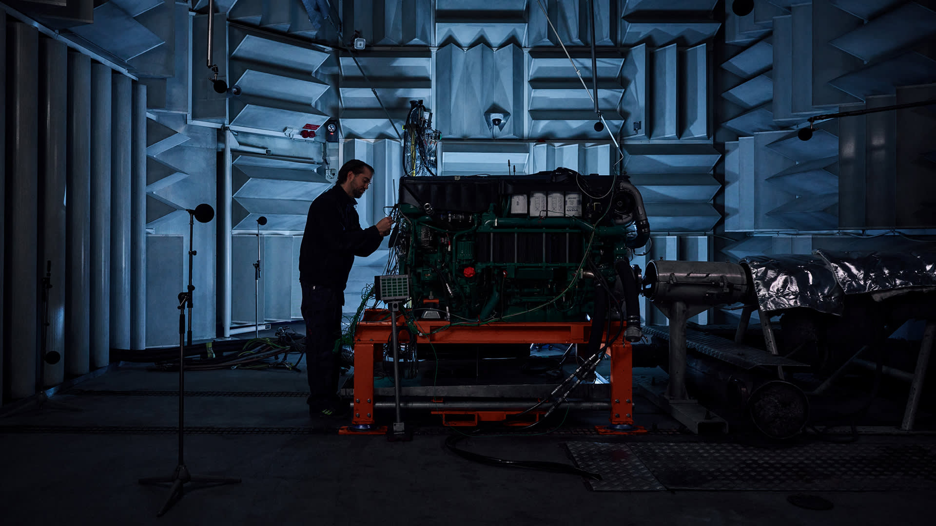 A person works on a large engine inside an acoustically treated room with soundproofing panels and recording equipment, illuminated by dim blue lighting.