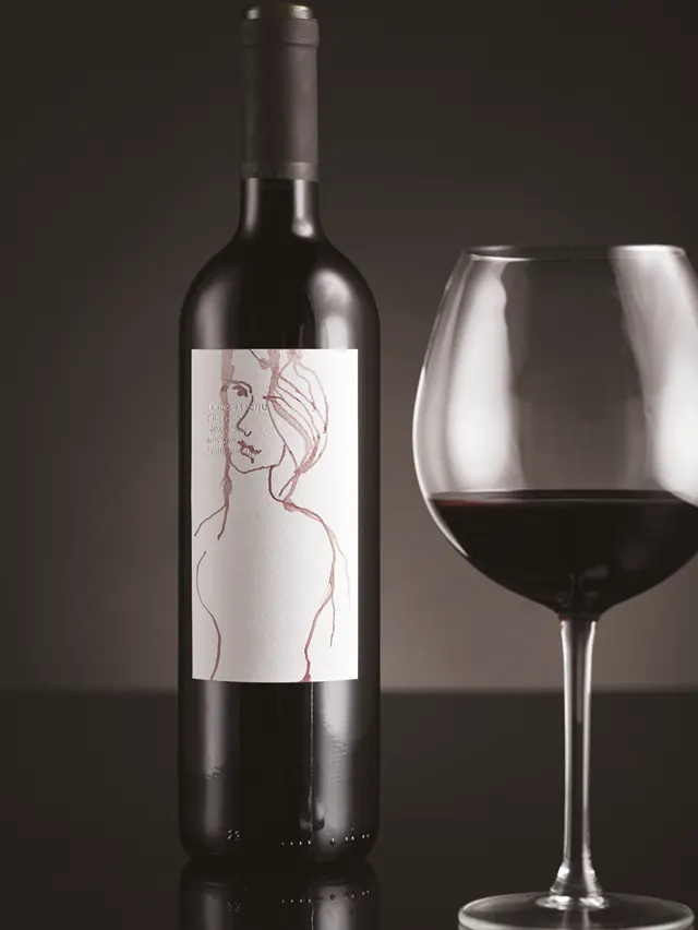 A bottle of red wine with a minimalist line drawing of a womans figure on the label stands next to a glass of red wine, both placed on a reflective surface against a dark background.