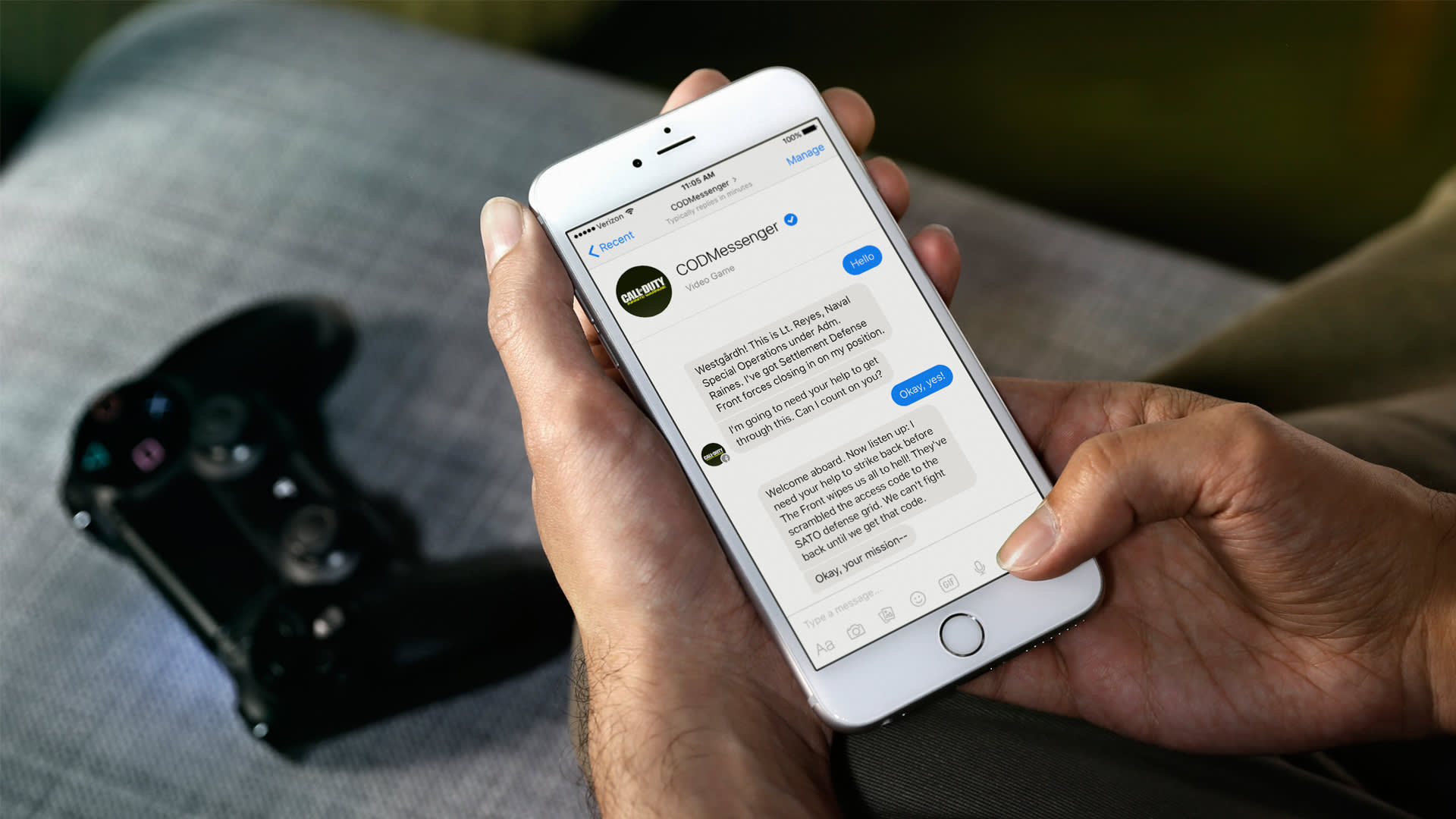 A person holds a smartphone displaying a Facebook Messenger chat with CDM messages, while sitting on a couch. A black video game controller rests on the gray fabric beside them.