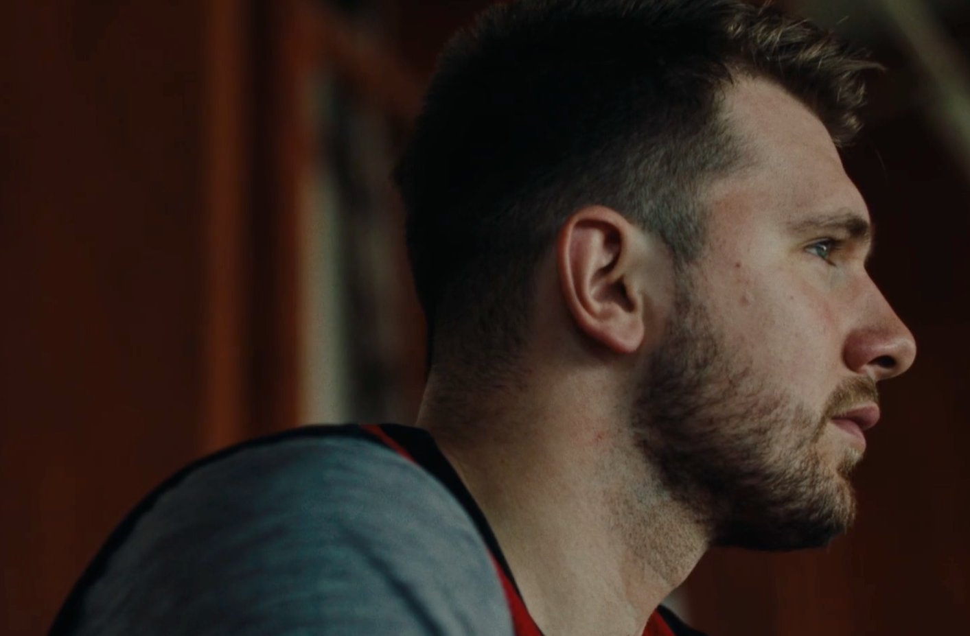 A close-up side profile of a man with short brown hair and a trimmed beard, wearing a gray shirt with a hint of red. He is looking thoughtfully into the distance, with a blurred brown background.