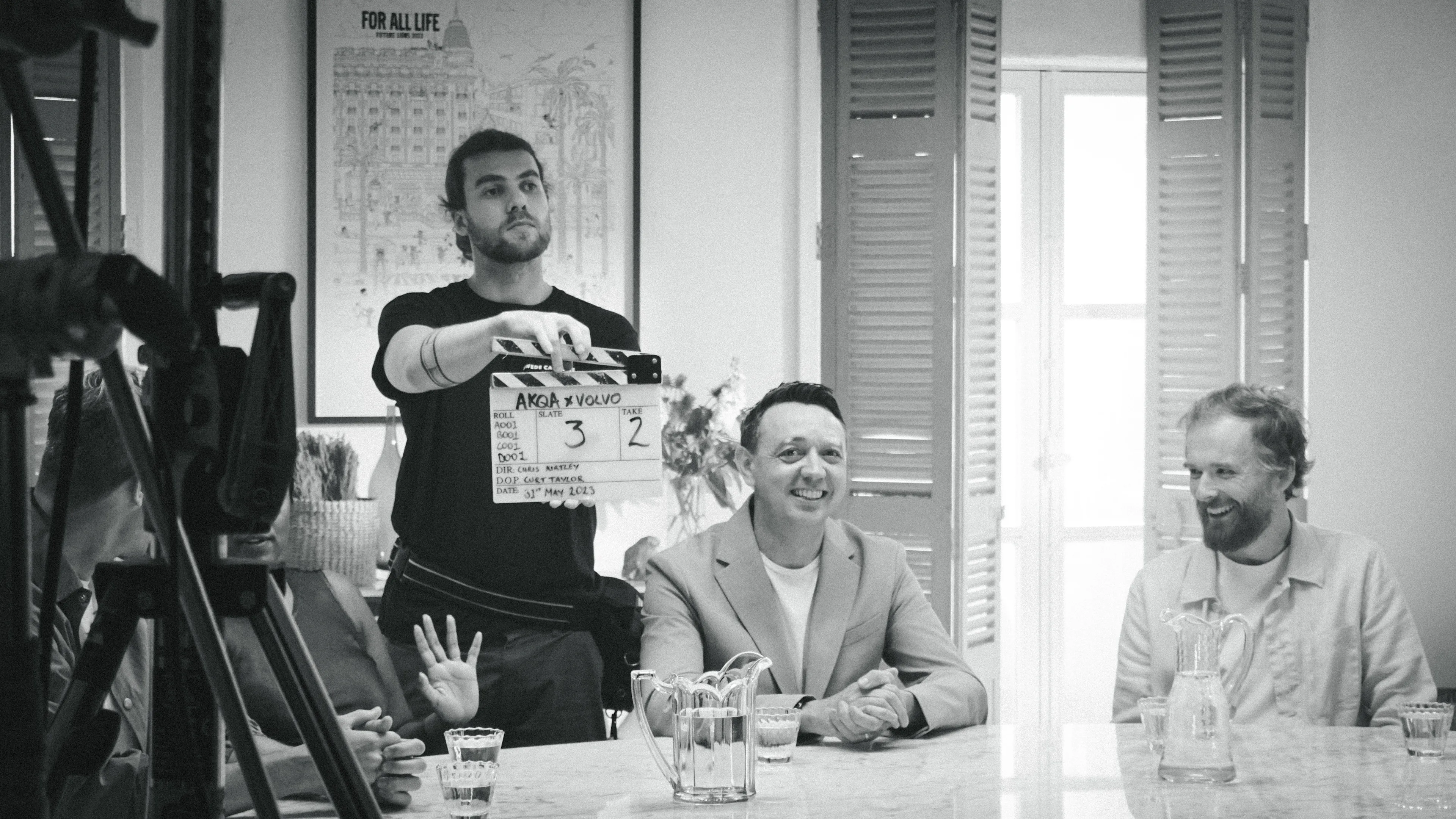 A black-and-white photo shows people seated at a table, smiling, as a crew member holds a clapperboard in front of them, suggesting a film or video production scene. Light streams in from a window behind them.