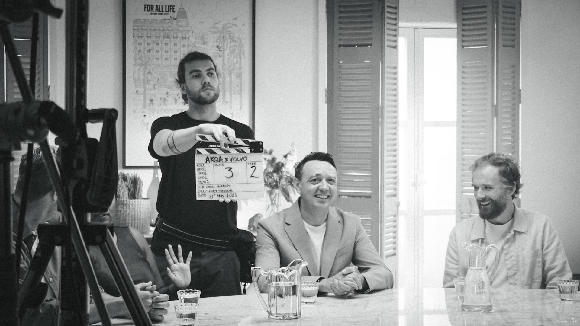 A black-and-white photo shows people seated at a table, smiling, as a crew member holds a clapperboard in front of them, suggesting a film or video production scene. Light streams in from a window behind them.