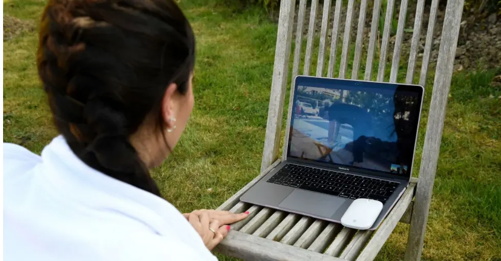 A woman with dark braided hair sits outside on a wooden chair, looking at a laptop screen showing a video call. A white computer mouse rests on the chair next to the laptop. Grass and garden are visible in the background.