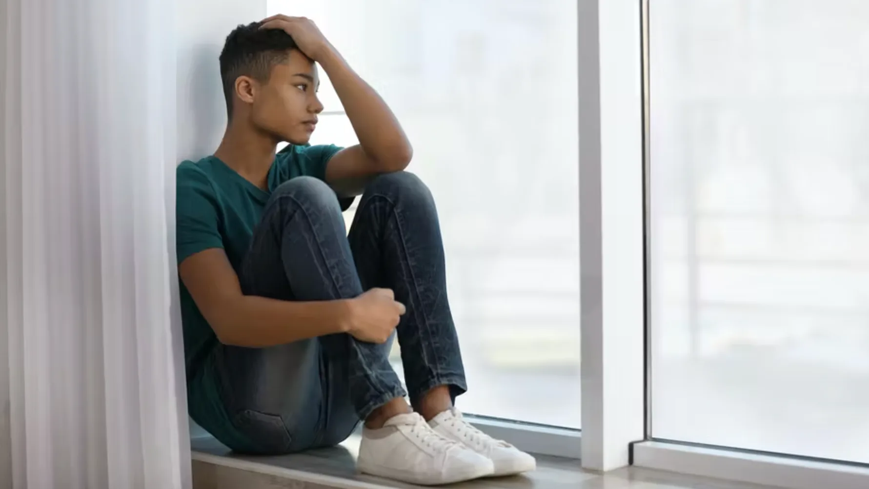 A young person sits on a windowsill with knees pulled to their chest, resting one hand on their head and looking outside with a thoughtful, pensive expression.