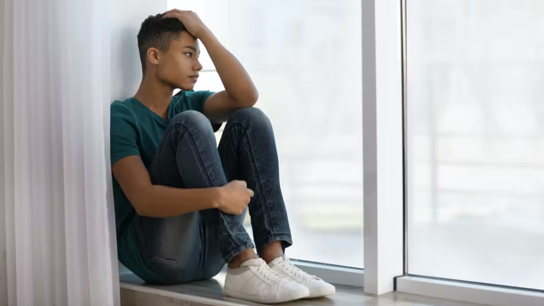 A young person sits on a windowsill with knees pulled to their chest, resting one hand on their head and looking outside with a thoughtful, pensive expression.