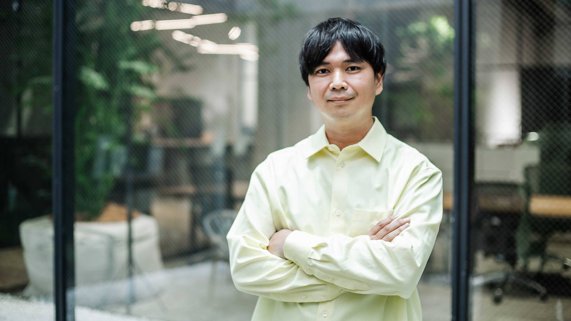 A man with dark hair wearing a light yellow shirt stands with arms crossed, smiling in a modern office with glass walls and greenery in the background.