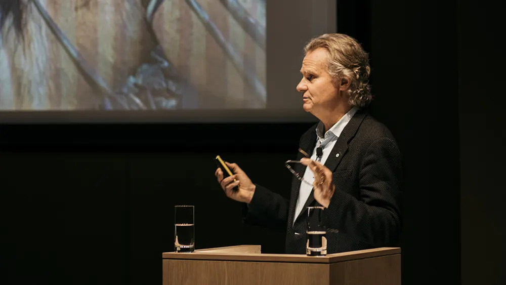 A man in a dark suit stands at a podium giving a presentation, holding glasses and a remote. Two glasses of water are on the podium, and a large projected image is visible behind him.
