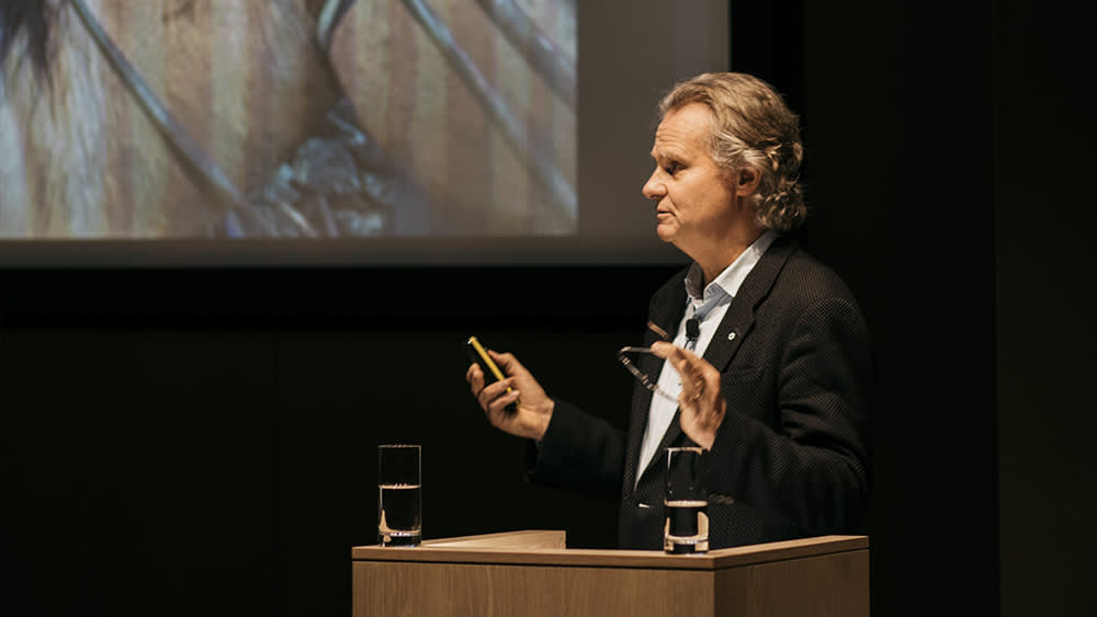 A man in a dark suit stands at a podium giving a presentation, holding glasses and a remote. Two glasses of water are on the podium, and a large projected image is visible behind him.