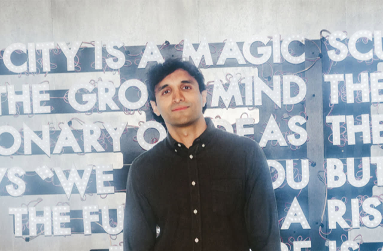 A person with short dark hair, wearing a dark button-up shirt, stands in front of a wall with illuminated, partially obscured text in white capital letters.
