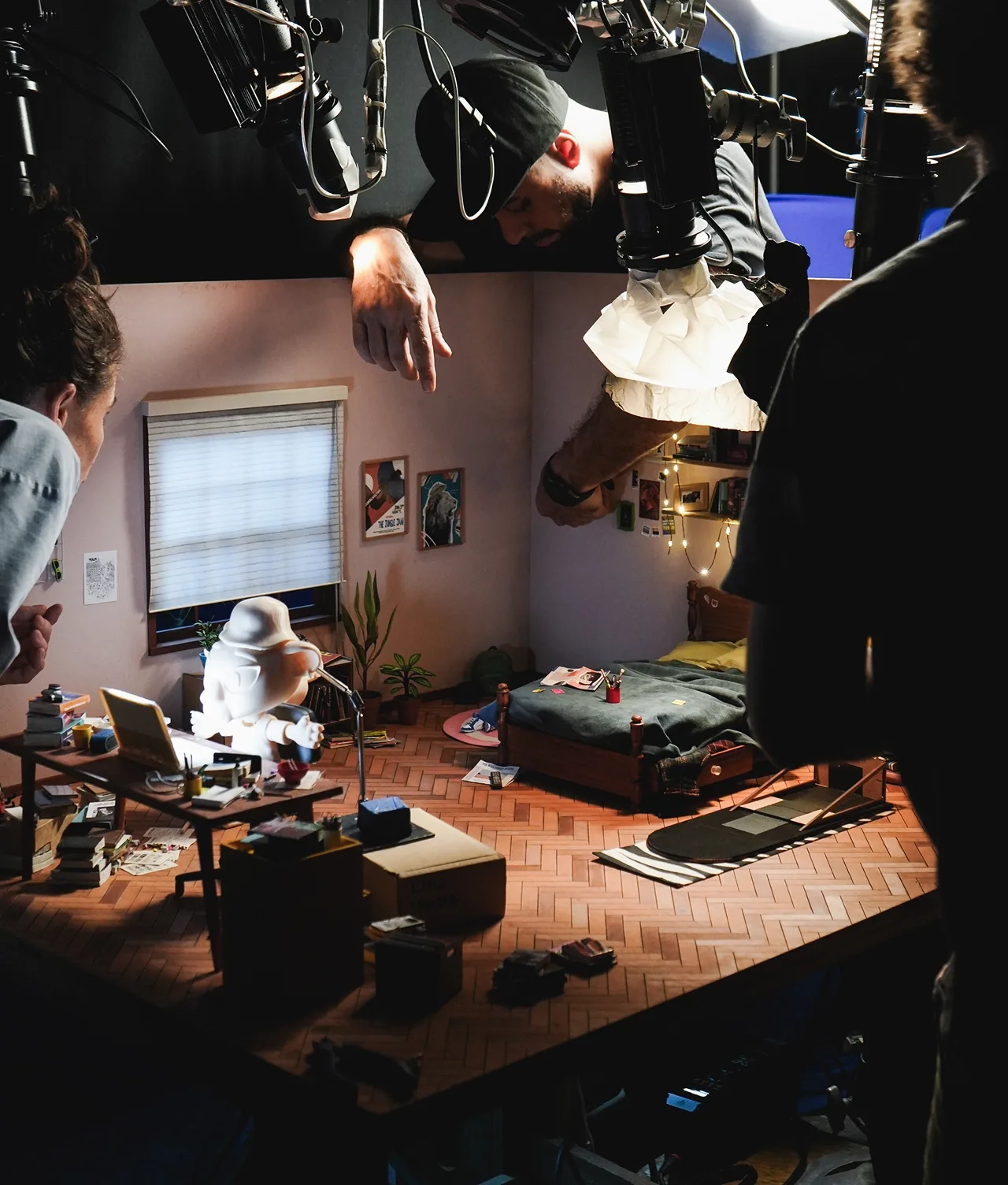 Several people arrange lighting and props around a detailed miniature bedroom set, with visible hands adjusting items atop the model, suggesting a stop-motion animation production in progress.