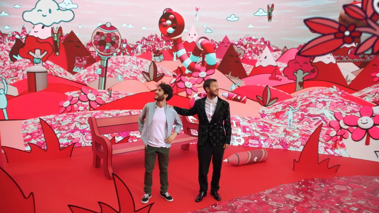 Two men stand in a vibrant, whimsical, candy-themed landscape with oversized sweets, a pink bench, and animated scenery in shades of red, pink, and white. One man gestures toward the colorful backdrop.