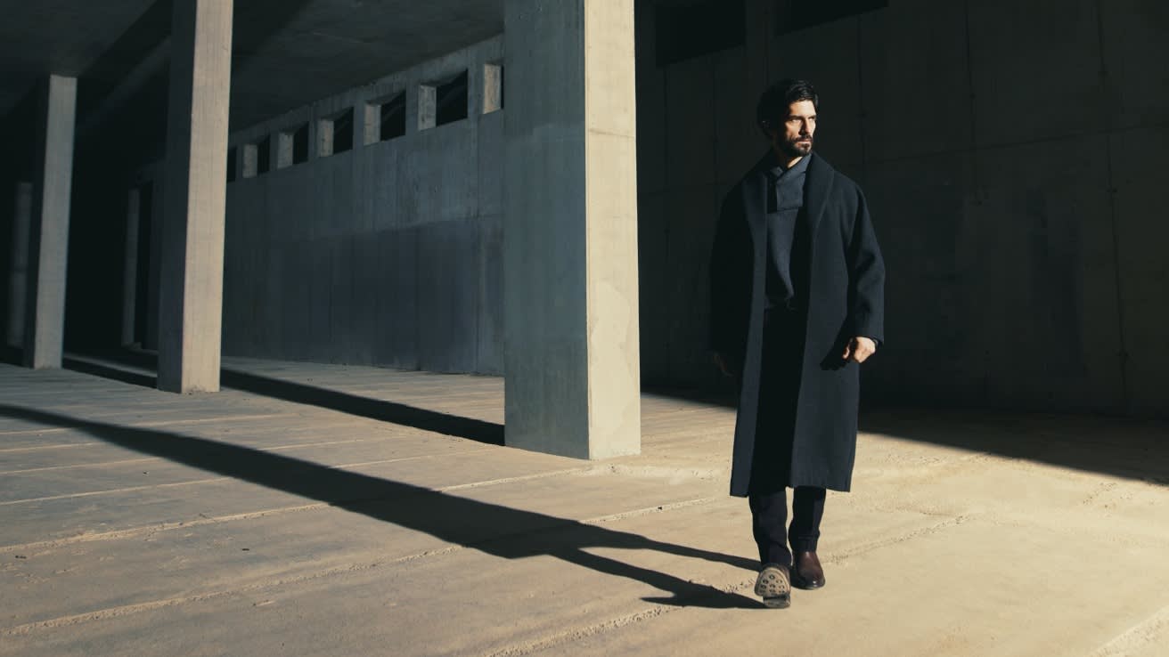 A man in a long dark coat walks through a modern, unfinished concrete building, casting a long shadow on the sunlit floor. He looks to the side and appears calm and thoughtful.