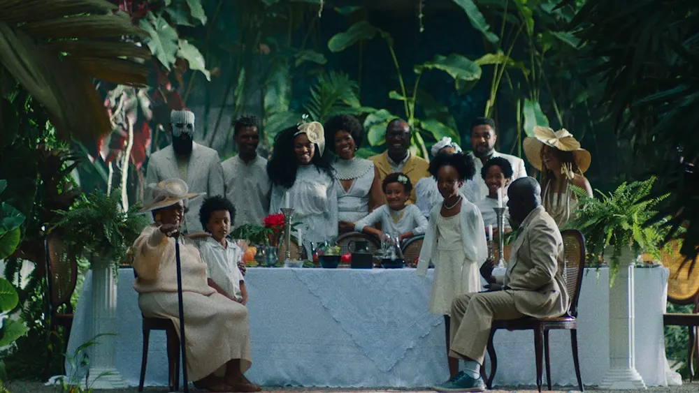 A group of people dressed in white and beige sit and stand around a table set for a meal in a lush, green garden. The scene is elegant and serene, with large leaves and plants surrounding them.