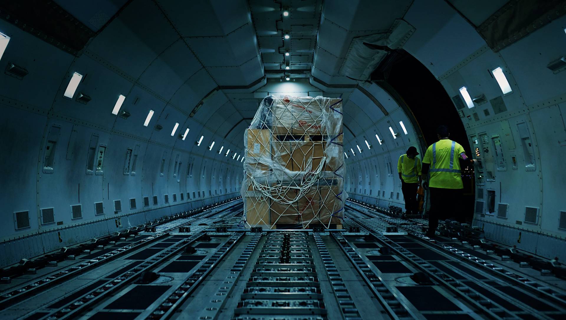 Large cargo boxes secured with netting sit inside the empty hold of a cargo plane, with two workers in yellow vests visible near the open rear of the aircraft.