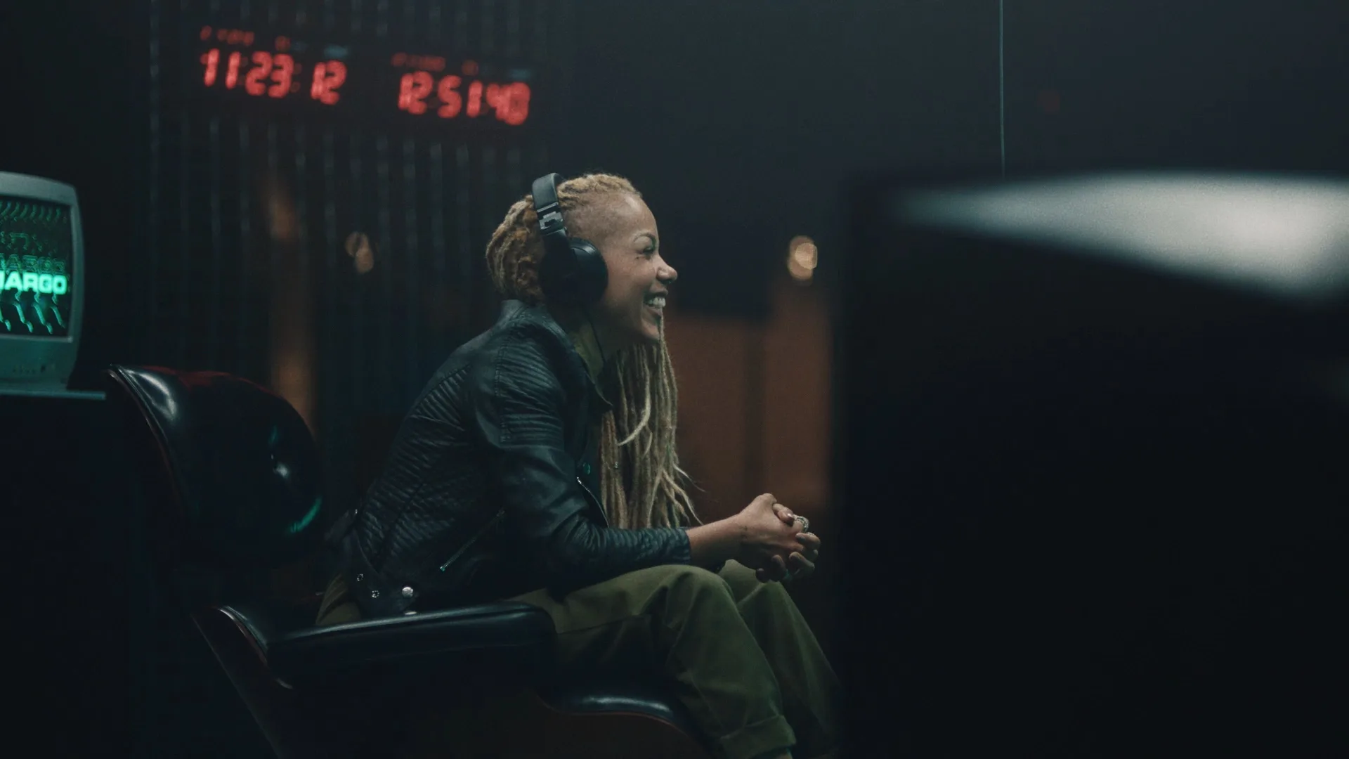 A person with long, light-colored braids sits on a chair, smiling and wearing headphones, in a dimly lit room with digital clocks displaying red numbers in the background.