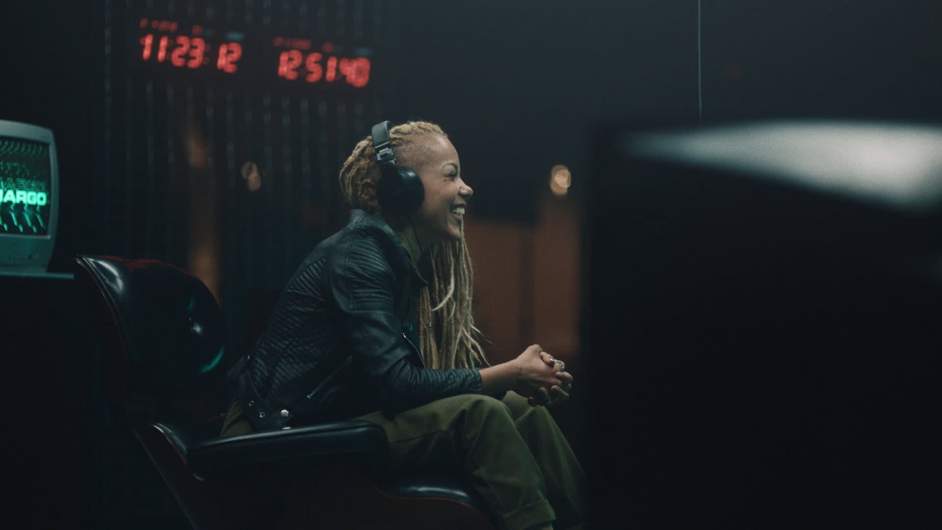 A person with long, light-colored braids sits on a chair, smiling and wearing headphones, in a dimly lit room with digital clocks displaying red numbers in the background.