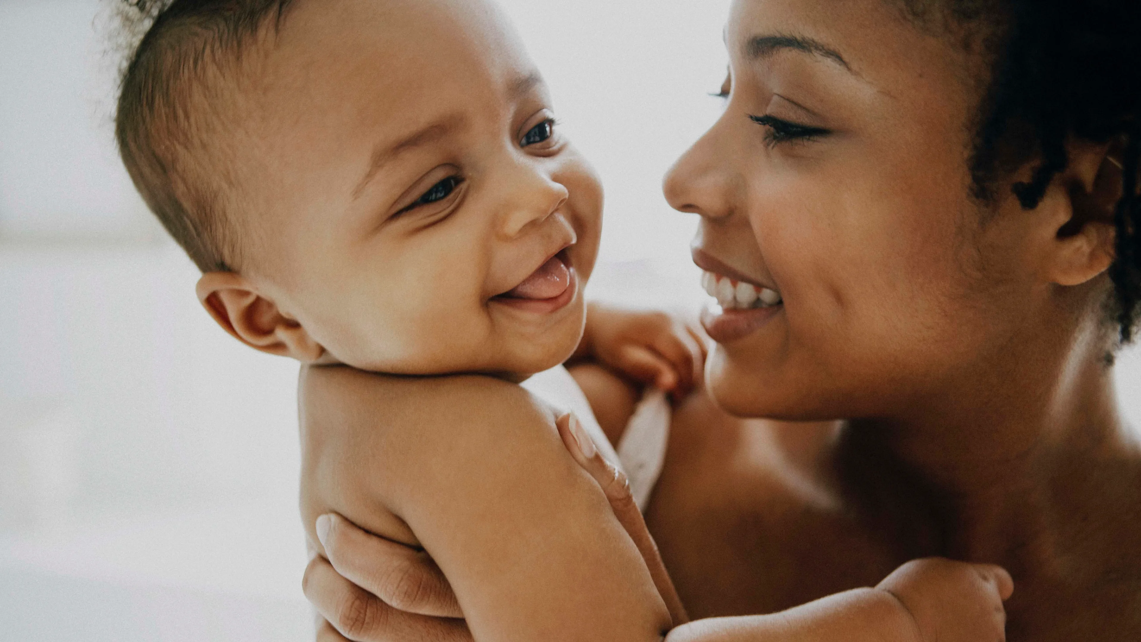 A smiling woman holds a happy baby who is looking at her and sticking out their tongue. Both appear joyful and are facing each other closely in a tender, bright setting.